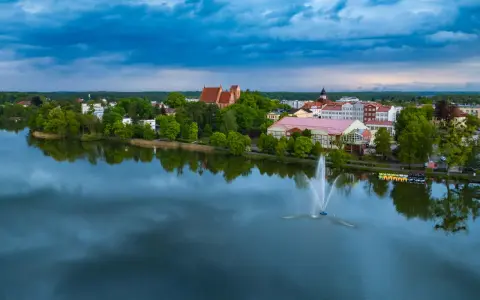 Widok na jezioro z fontanną i zabudowaniami, w tym kościołem. Odkryj iława atrakcje, które zachwycają o każdej porze dnia.