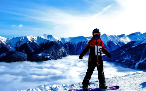 Snowboardzista w kurtce hardshell stoi na szczycie góry, nad morzem chmur. Góry w tle.