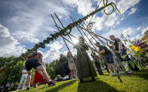Midsommarstången reses av en grupp människor, en traditionell del av hur firar man midsommar. Solen skiner genom molnen.