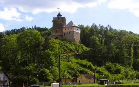 Zamek w Czchowie góruje nad zielonym wzgórzem, otoczony drzewami. Na szczycie wieży powiewa flaga.