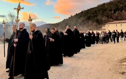 Monks in black robes process with a cross and lanterns, their solemn religious chants echoing as they walk towards a building at sunset.