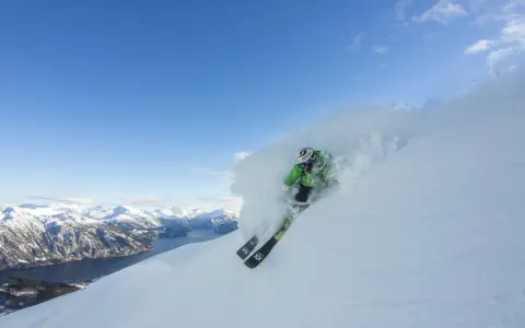 En skidåkare i grön jacka plöjer genom pudersnö med en fantastisk fjordvy i bakgrunden. Stranda ski-skidorna skär genom snön.