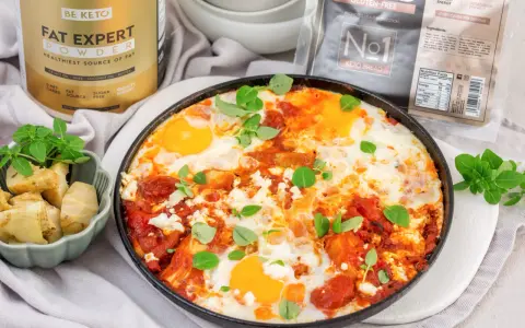 Pyszne śniadanie low carb: jajka sadzone w sosie pomidorowym z fetą i ziołami, obok pieczone karczochy. Obok produkty BE KETO.