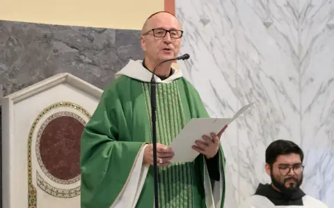 A man with a Benedictine title, wearing a green chasuble, speaks into a microphone while holding a paper.