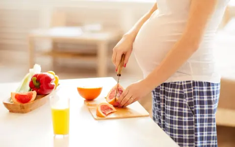 Schwanger, die Grapefruit für einen gesunden Snack schneidet. Nebenan liegen Paprika und ein Glas Saft.