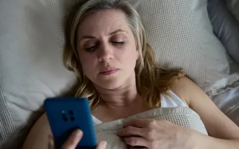 Kobieta leży w łóżku, trzymając telefon. Może to być związane z problemem oddawania moczu w nocy jeden raz.