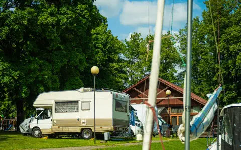 Słoneczny dzień na kempingu na Mazurach. Kamper stoi na trawie obok drewnianego budynku, w tle zielone drzewa i plac zabaw.
