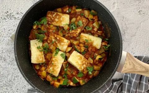 Smażony ser halloumi z warzywami w sosie pomidorowym, posypany natką pietruszki, w czarnej patelni.