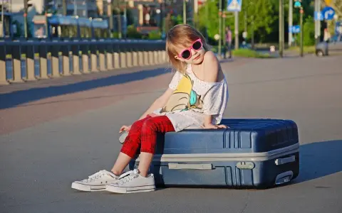 Dziewczynka w okularach siedzi na walizce, gotowa na wyjazd dziecka za granicę.