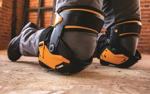 Protection des genoux pour éviter toute maladie professionnelle. Des genouillères robustes et orange protègent les genoux d'un travailleur sur un sol poussiéreux.