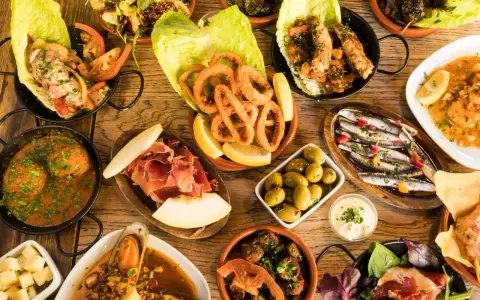 Tapas selber machen: Eine Auswahl an köstlichen spanischen Kleinigkeiten wie frittierte Tintenfischringe, Sardinen, Oliven und Schinken auf einem Holztisch.