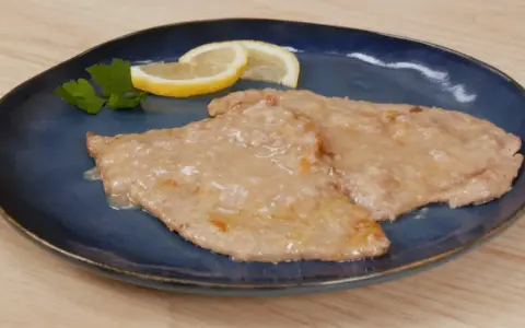 Due scaloppine al limone cremose, guarnite con prezzemolo e spicchi di limone, su un piatto blu.