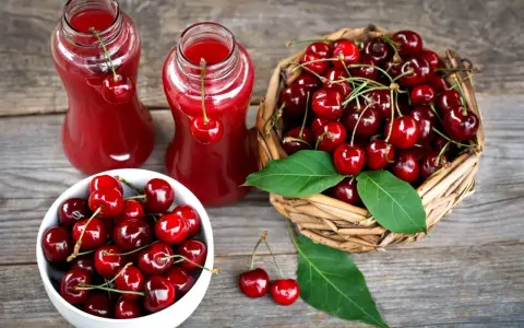 Dwa szklane dzbanki z rubinowym sokiem z czereśni, obok koszyk pełen świeżych owoców i biała miseczka z czereśniami.