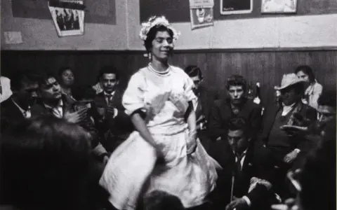 Una mujer con corona de flores baila en una celebración, rodeada de gente. Podría ser una boda gitana.