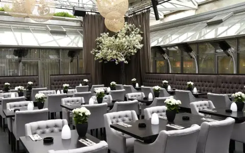 Elegantes Restaurant mit grauen Sitzmöbeln und dunklen Tischen, dekoriert mit weißen Blumen. Ein großer Kirschblütenbaum im Zentrum, beleuchtet von kugelförmigen Lampen. Perfekt für ein Rendezvous, wie ein Oscar-Moment im Apropos.