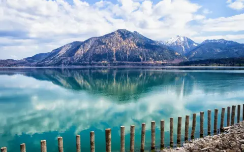 Spiegelglatter See mit Bergen im Hintergrund, wie man ihn in Bayern sehen kann. Holzpfähle ragen aus dem türkisfarbenen Wasser.