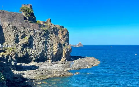 Castello Normanno a Aci Trezza, un luogo da vedere. Roccioso promontorio sul mare blu, con un antico castello in pietra e vegetazione.