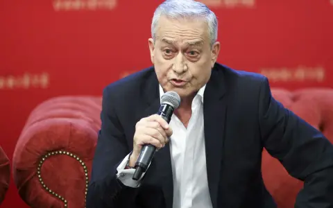 Janusz Szydłowski przemawia do mikrofonu, siedząc na czerwonej, pikowanej kanapie.