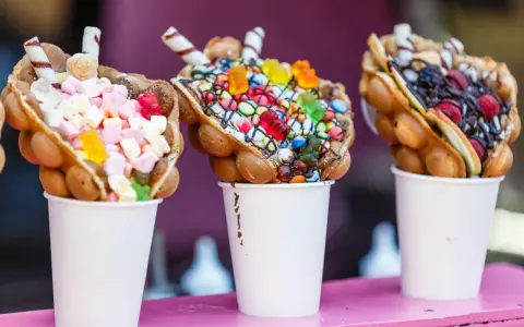 Trois bubble waffles colorés, garnis de bonbons, guimauves et fruits, présentés dans des gobelets blancs.