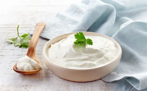 Gęsta, biała śmietana 30 kcal w miseczce, obok drewniana łyżka z porcją. Idealna do deserów i sosów.