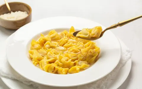 Un cucchiaio d'oro solleva un boccone di cappelletti in brodo fumante da un piatto bianco.