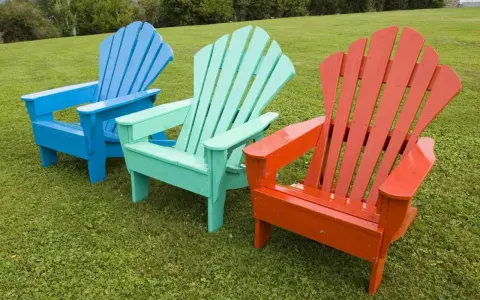 Tres sillas de madera de estilo Adirondack, pintadas de azul, verde menta y naranja, lucen vibrantes sobre el césped. ¡Un proyecto de pintar sillas de madera sin lijar que da vida al jardín!