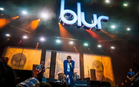 Medlemmar av Blur uppträder på scenen under en konsert, med den ikoniska logotypen lysande ovanför.