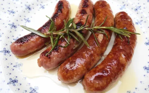 Salchichas al vino, doradas y jugosas, reposan sobre un plato blanco con delicados motivos azules, adornadas con ramitas de romero fresco.