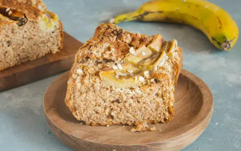 Pyszny chlebek bananowy fit z płatkami owsianymi na wierzchu, podany na drewnianym talerzyku. Obok leży dojrzały banan.