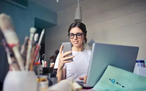 Kobieta w okularach do komputera przegląda telefon, siedząc przy laptopie.