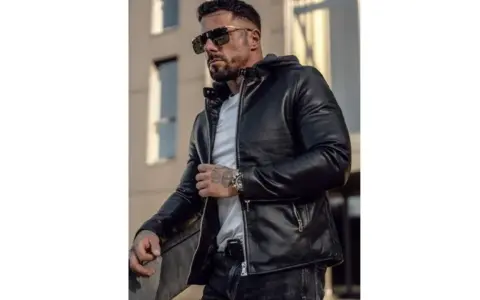 Mężczyzna w stylowej, czarnej kurtce woskowanej męskiej, z kapturem, okularach przeciwsłonecznych i tatuażami.