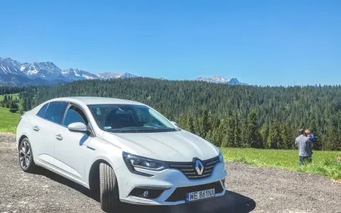 Biały Renault Megane Sedan zaparkowany na szutrowej drodze z widokiem na góry i las. Na trawie stoi mężczyzna z dzieckiem.