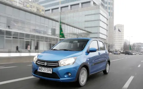 Niebieski Suzuki Celerio jedzie ulicą w mieście, mijając nowoczesne budynki i innych uczestników ruchu.