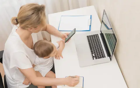 Kobieta z dzieckiem na kolanach sprawdza na telefonie informacje o zasiłku macierzyńskim 80 czy 100% i 60. Obok laptop i notatnik.