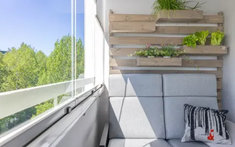 Przytulny balkon w bloku z szarą kanapą, poduszką z liskiem i zielenią na drewnianej ściance.