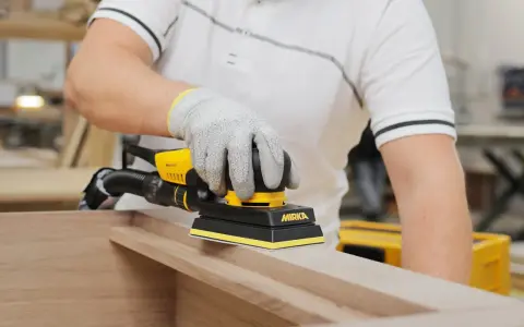 Un artisan utilise une ponceuse Mirka pour comment poncer un meuble avec des moulures. La ponceuse est jaune et noire, et l'artisan porte des gants de protection.