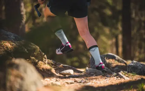 Beine einer Person beim **trail running** auf einem steinigen Waldweg. Leichte Sportschuhe und helle Socken mit Logo.