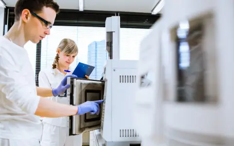 W laboratorium młoda para naukowców pracuje przy aparaturze. On w okularach, ona z notatkami.