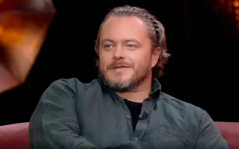 Tomasz Schuchardt, znany z wielu filmów, seriali i programów, z charakterystyczną fryzurą i niebieskimi oczami, siedzi na kanapie.