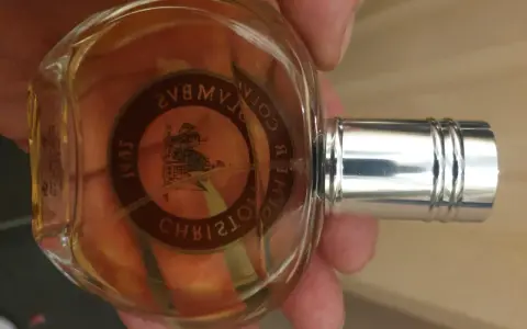 Szklana butelka z bursztynowym płynem, ozdobiona okrągłą etykietą z wizerunkiem statku i napisem "Christopher Columbus perfumy".