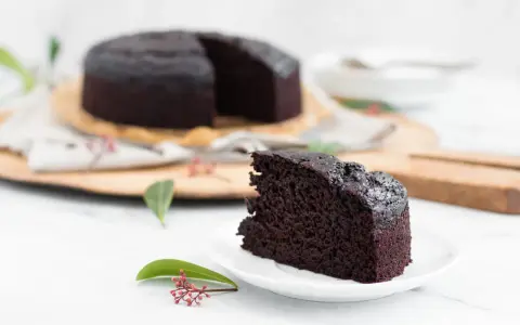 Ein saftiges Stück veganer Schokokuchen liegt auf einem Teller. Im Hintergrund ein ganzer Kuchen, dekoriert mit grünen Blättern und roten Beeren.