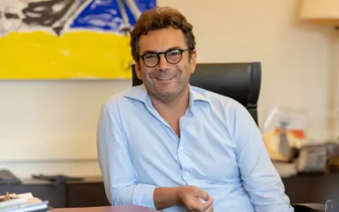 A man with glasses and a light blue shirt smiles while sitting at a desk with abstract art in the background.