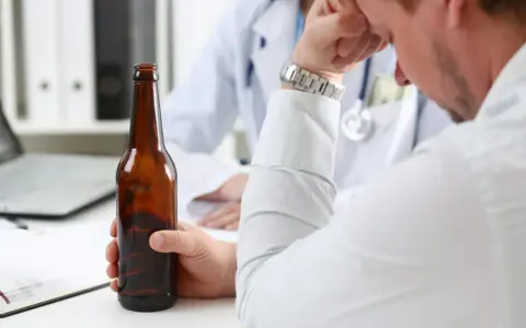 Mężczyzna trzyma butelkę piwa, siedząc przed lekarzem. Rozmowa sugeruje skierowanie na detoks alkoholowy.