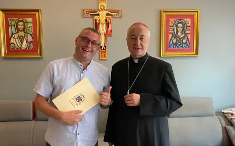 Uśmiechnięty mężczyzna z dokumentem i biskup pozują na tle krzyża i obrazów. Mężczyzna trzyma dokument z herbem, a biskup ma na sobie sutannę.