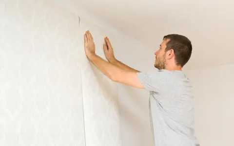 Hombre colocando papel pintado con patrón floral en una pared. Aprende como poner papel pintado tú mismo.