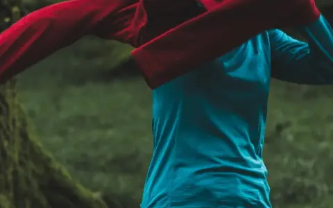 Person zieht ein rotes Oberteil über ein blaues Funktionsshirt. Das blaue Shirt ist ein Baselayer, der die Körperwärme reguliert.
