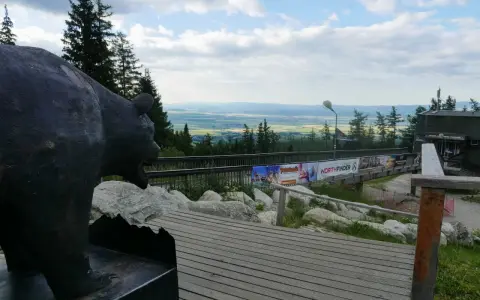 Pomnik niedźwiedzia na tle panoramy Tatr poza szlakiem. Drewniana kładka prowadzi do budynku z widokiem na dolinę.