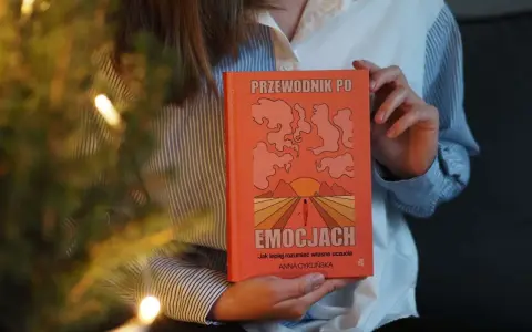 Osoba trzyma pomarańczową książkę "Przewodnik po emocjach". Okładka przedstawia abstrakcyjne kształty i zachód słońca.