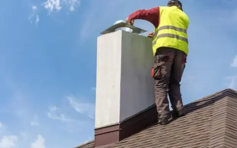 Pracownik na dachu montuje przedłużenie komina. Niebo błękitne z chmurkami.