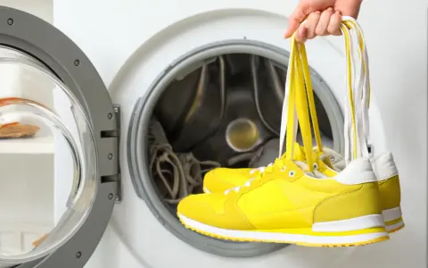 Czy buty zamszowe można prać w pralce? Ręka trzyma żółte sneakersy nad otwartym bębnem pralki.